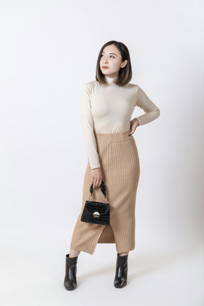 Elegant studio shot of a fashionable Asian woman in a beige turtleneck and skirt with a black handbag.